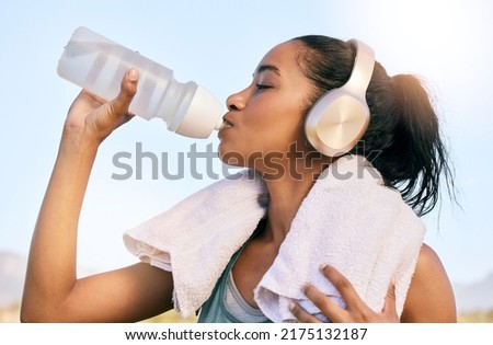 An active fit woman wearing wireless headphones drinking water from a bottle after exercising outdoors. Female athlete quenching thirst and cooling down with a towel after training workout outside