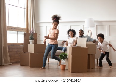 Active Funny Kids Play Run Around Cardboard Boxes Having Fun On Moving Day With Black Parents Sit On Sofa, Happy African American Family Children Mom Dad Laughing In Living Room Celebrate Relocation