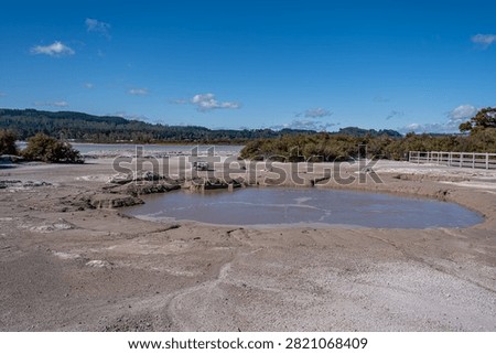Active geothermal mud pool surrounded by cracked earth and native thermal vegetation including Manuka (Leptospermum scoparium) at Sulphur Point on the shore of Lake Rotorua, Rotorua, Bay of Plenty, New Zealand. Accessible wooden boardwalk walkway on the right, bright sunstar flare radiating in a vivid blue sky, with high contrast daylight, wide angle perspective, and scenic natural thermal landscape.