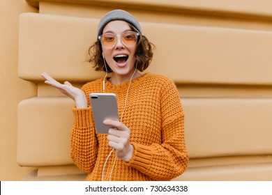 Active girl in trendy cozy attire posing with happy face expression, holding smartphone. Excited young lady in knitted outfit waving hand in front of building after phone call.