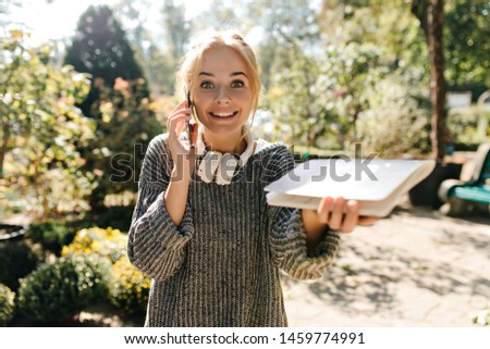 Active green-eyed girl looks at camera with pleasant surprise, talking on phone and showing off her notebook