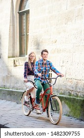 Active Happy Tourist Couple, Bearded Man And Smiling Long-haired Blond Woman In White Skirt And Glasses Cycling Tandem Bike Along Paved City Sidewalk By Old Ancient Stone Building.