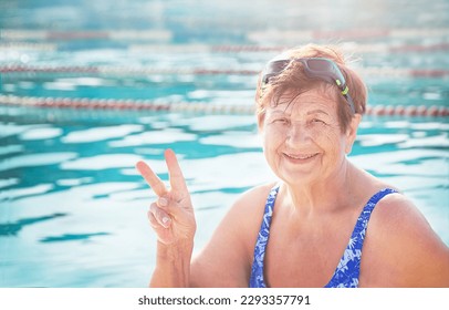 Active, healthy happy senior (elderly) woman (over age of 50) in swimsuit, sport goggles smiling with happy face doing victory sign (number two) in sunny summer day. Swimming pool background. - Powered by Shutterstock