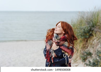 Active Healthy Young Woman Enjoying A Sandy Tropical Winter Beach Standing In A Warm Anorak And Scarf Looking Up Into The Sun With A Blissful Smile And Closed Eyes