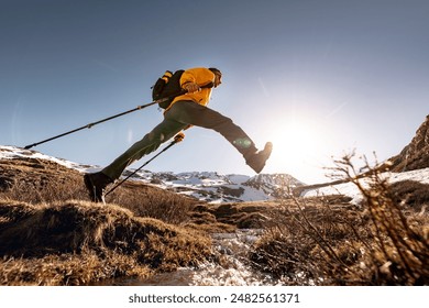 Active hiker tourist in bright yellow jacket with backpack and hiking poles make long jump over small stream against blue sky and sun - Powered by Shutterstock