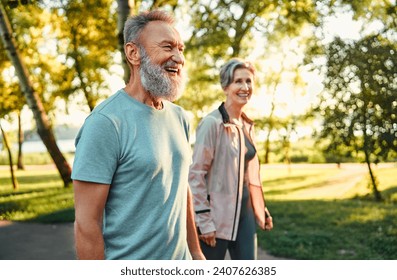 Active hobby on retirement. Grey haired caucasian man with beautiful wife strolling at summer park and smiling cheerfully. Fitness retired couple in sport attire taking break after morning run. - Powered by Shutterstock