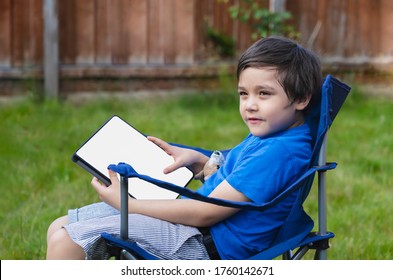 Active Kid Sitting On Chair In The Garden Playing Game Or Watching Cartoons On Tablet, Handsome Young Boy Relaxing In The Garden On Sunny Day Summer, New Normal Lifestyle Concept