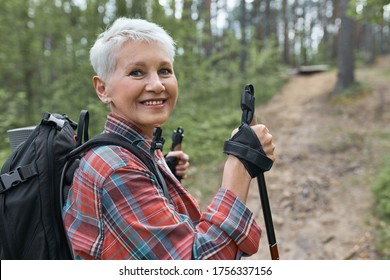 Active Lifestyle, Aging, Travel And Hiking Concept. Outdoor Image Of Beautiful Energetic Mature Woman With Backpack Using Poles, Enjoying Nordic Walking In Forest, Looking At Camera With Happy Smile