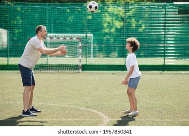 Active Lifestyle Concept. Side View Of Sportive Single Adult Dad Playing Football With His Son On Green Grass At Soccer Pitch, Throwing The Ball To Boy, Family Spending Time Together. Recreation