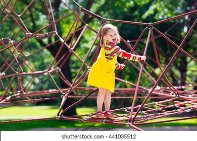 Active little child playing on climbing net and jumping on trampoline at school yard playground. Kids play and climb outdoors on sunny summer day. Cute girl on nest swing at preschool sport center.