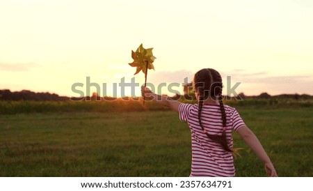 Active little girl runs playing with windmill along meadow lit by sunset light