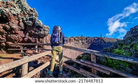 Active Man with backpack standing on fence at the edge of active volcano crater of Mount Vesuvius, Province of Naples, Campania, Italy, Europe, EU. Person enjoying the view on the mountain summit. Awe