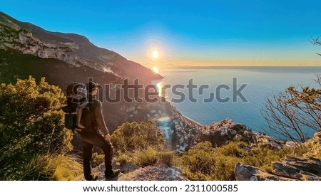 Active man with backpack watching sunrise from hiking trail Path of Gods between Positano and Praiano, Amalfi Coast, Campania, Italy, Europe. Calm water surface reflecting sun ray at Mediterranean Sea