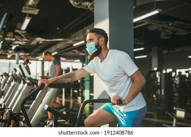 An Active Man In A Protective Medical Mask Training In Gym, Riding A Stationary Exercise Bike. Sport And Quarantine.