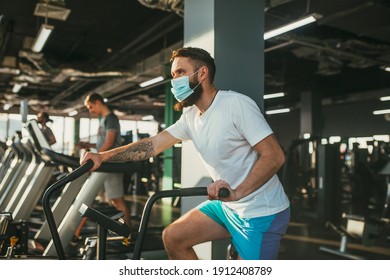 An Active Man In A Protective Medical Mask Training In Gym, Riding A Stationary Exercise Bike. Sport And Quarantine.