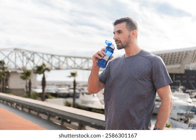 Active Man Quenching Thirst With Refreshing Water During Intense Workout Session In Comfortable Sportwear On A Sunny Day By The Marina Near The Boats And Bridges In A Beautiful Coastal Area