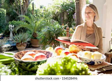 Active Mature Attractive Woman Sitting In Her Home Garden Reading A Cook Book Next To A Table Full Of Organic Vegetables And Fruit In Abundance, During A Sunny Day Outdoors.