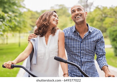 Active mature couple riding bikes during summer vacation. Happy couple cycling and smiling. Cheerful multiethnic couple loving and looking away. Mature woman in love looking her boyfriend. 