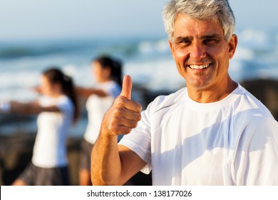 active mature man giving thumb up on beach with family exercising on background - Powered by Shutterstock