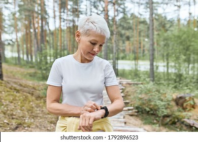 Active Mature Woman With Short Blonde Hair Posing Outdoors, Getting Ready For Jogging Exercise, Setting Smart Watch, Tracking Heart Rate And Pulse. People, Sports, Fitness And Technology Concept