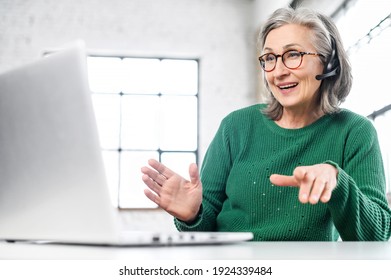 Active mature woman using a laptop for remote work from the home office. Smiling elderly lady wearing wireless headset talking on a video conference, has a video meeting. Senior teacher leads webinar