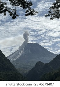 Active Merapi volcano emitting a plume of smoke and ash into the sky, surrounded by lush green hills and a dramatic cloudy sky. 