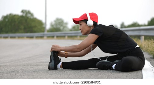 Active middle aged Asian sporty woman in black sportswear with headphones and baseball cap stretching her leg warm up after jogging on dam ridge, female fitness athlete exercising outdoors - Powered by Shutterstock