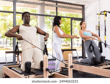 Active middle-aged African American man and young Woman doing pilates exercises on pilates workout machines at gym, instructor helping and controlling - Powered by Shutterstock