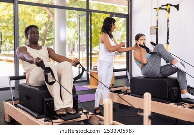 Active middle-aged African American man and young Woman doing pilates exercises on pilates workout machines at gym, instructor helping and controlling - Powered by Shutterstock