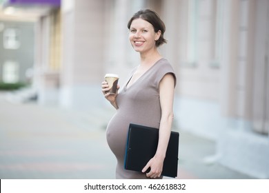 Active Mom. Portrait Of Happy Young Business Pregnant Woman Looking At Camera While Standing On The Street With Document Folder. Smiling Future Mom Working On Her Late Pregnancy Period