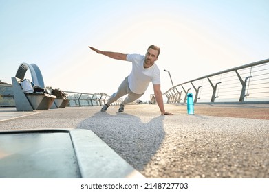 Active Muscular European Middle Aged Man, Athlete, Sportsman Doing Push-ups From The Floor, Alternately Raising One Arm During An Outdoor Workout At Dawn On The City Bridge