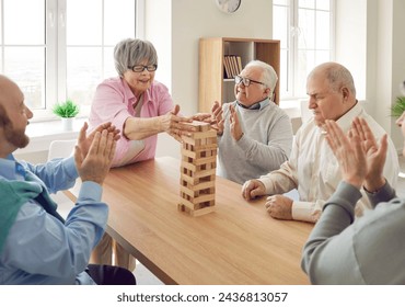 Active old age. Group of senior people sitting at table in nursing home and playing jenga together. Cheerful retired men applauding mature woman who successfully pulls out wooden block in board game. - Powered by Shutterstock