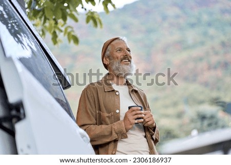 Active old happy hipster man standing near rv camper van on vacation. Mature traveler looking away enjoying view, holding drinking coffee waking up in the morning in camping tourism nature park.