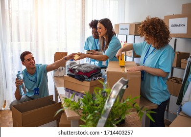 Active People Enjoys Volunteering At Food Bank. Voluntary Company. Curly Beautiful Girl Working In Voluntary Company With Friends Putting Food For Donation Into Boxes. 