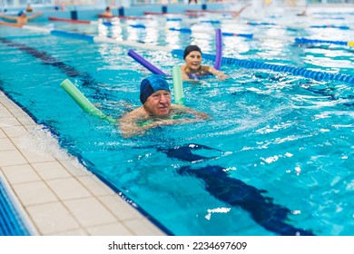 Active positive caucasian married senior adult couple swimming in a sport pool. People in swimming caps using colourful foam pool noodles for support. High quality photo
