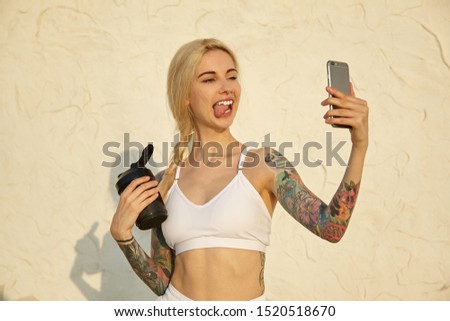 Active pretty young tattooed blonde woman wearing her hair in braid making photo of herself with smartphone over white wall, making funny face and showing tongue