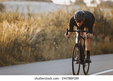 Active professional sportsman cyclist wearing black sports outfit, helmet and glasses riding bike at the paved road outdoors. Strong man improving skills and getting ready for cycling competition