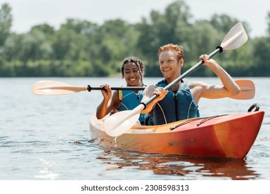 hombre de cabeza roja activo y encantadora mujer afroamericana en chalecos salvavidas que pasan tiempo en el río mientras navegan en kayak deportivo en un pintoresco lago sobre fondo borroso en verano