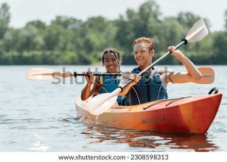 active redhead man and charming african american woman in life vests spending time on river while sailing in sportive kayak on picturesque lake on blurred background in summer