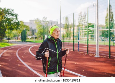 Active Rest Of The Elderly Theme. Sports And Health In Retirement. Caucasian Very Old Woman With Deep Wrinkles Doing Nordic Walking Exercises With Sticks Outside In The City Stadium.