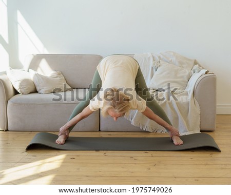 Active senior 60s woman in sports wear practicing downward facing dog yoga pose while exercising at home. Mature flexible lady doing wide legged forward bend or Prasarita Padottanasana posture
