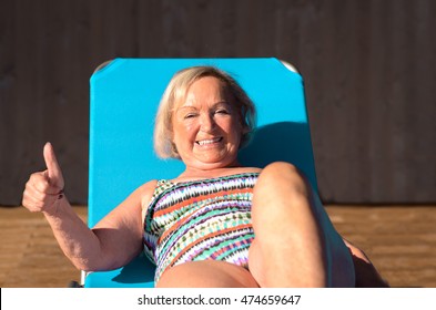 Active Senior Blond Woman Sunbathing On A Recliner Chair Giving A Thumbs Up Of Sign Of Approval With A Beaming Smile