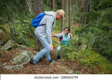 Active Senior Couple Hiking On The Forest Rock. Mature Man Helping Woman Climbing Up. Happily Smiling. Scenic View Green Wood. Healthy Lifestyle. Finland.