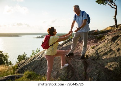 Active Senior Couple Hiking On The Top Of Rock. Mature Man Helping Woman Climbing Up. Happily Smiling. Scenic View Of Gulf And Sea. Healthy Lifestyle. Finland.