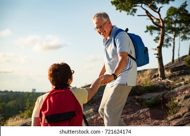 Active Senior Couple Hiking On The Top Of Rock. Mature Man Helping Woman Climbing Up. Happily Smiling. Scenic View Of Gulf And Sea. Healthy Lifestyle. Finland.