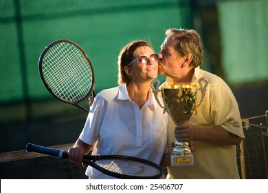 Active senior couple is posing on the tennis court with tennis racket and cup in hand. Outdoor, sunlight.