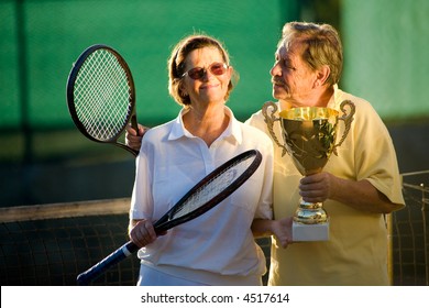Active senior couple is posing on the tennis court with tennis racket and cup in hand. Outdoor, sunlight.