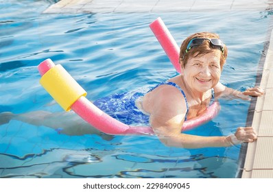 Active senior (elderly) woman (over age of 50) in sport goggles, swimsuit doing aqua fitness with swim noodles in swimming pool. Mature female smiling with happy face in summer day. Healthy lifestyle.