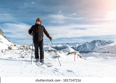 Active Senior Experienced Mature Old Person In Ski Helmet, Goggles And Black Suit Stand On Mountain Peak Enjoy Winter Extreme Sport Activities. Man On Stunning Panoramic Mountains Sunshine Background