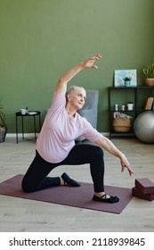 Active Senior Female In Black Leggins And Beige T-shirt Doing Physical Exercise On Yoga Mat Against Domestic Plants And Furniture In Living-room